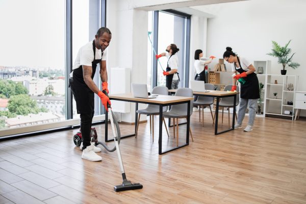 Professional service team of cleaning company. Young adult African American man wearing black apron and red rubber gloves vacuuming floor of modern light office.