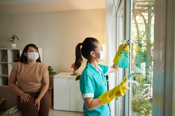 Young woman from cleaning service washing windows in the room with owner watching for process